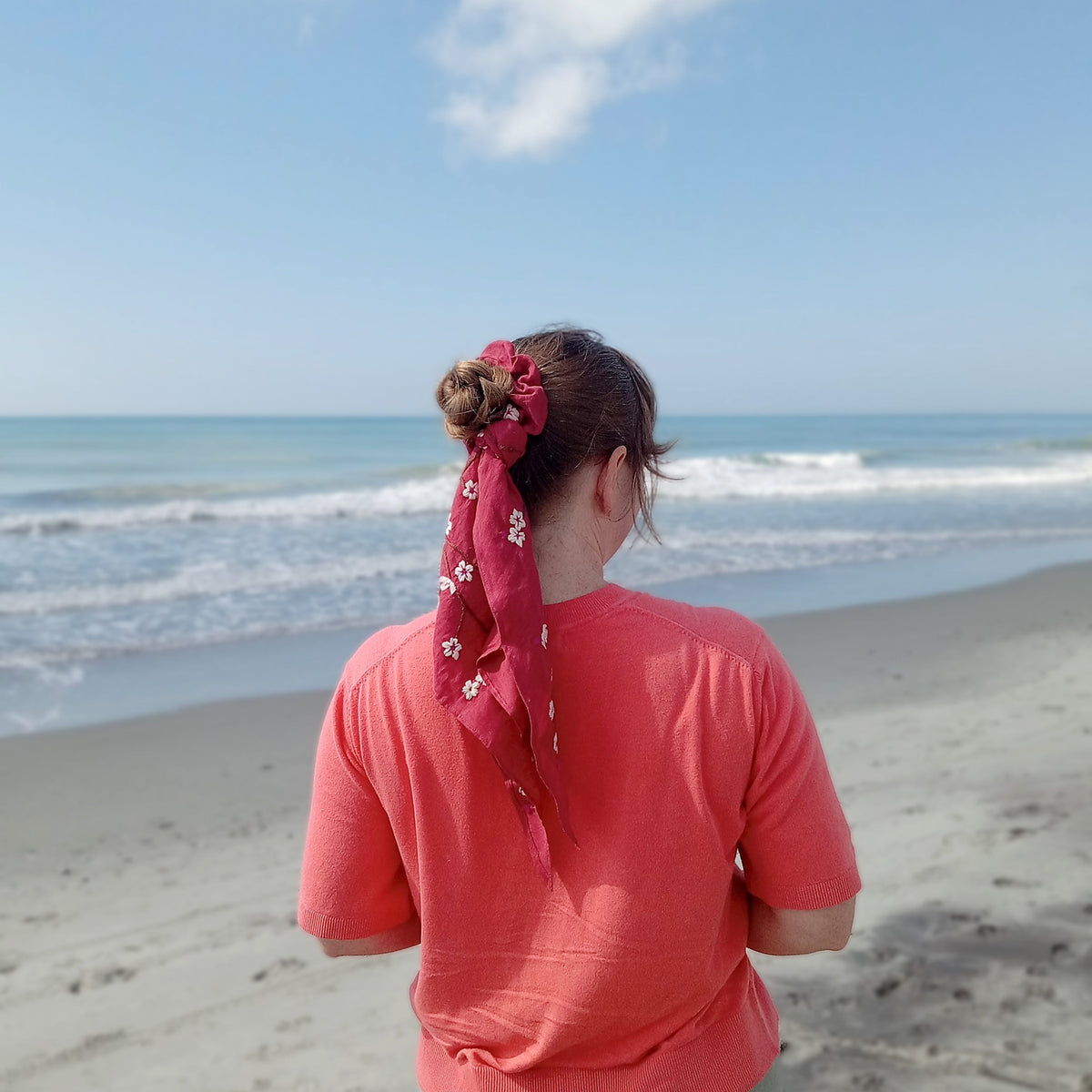 A woman standing on a sandy beach with the ocean and blue sky behind her, viewed from behind. She is wearing a coral pink t-shirt and light olive green pants, with a red hand-embroidered scarf tied in her hair.
