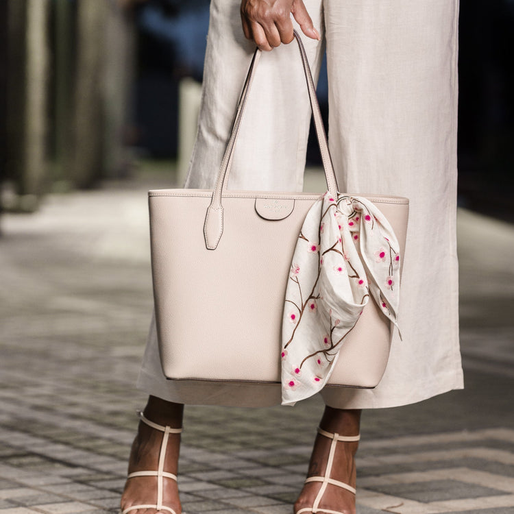 Person holding a beige handbag with a floral scarf, wearing beige pants and sandals on a blurred street background.
