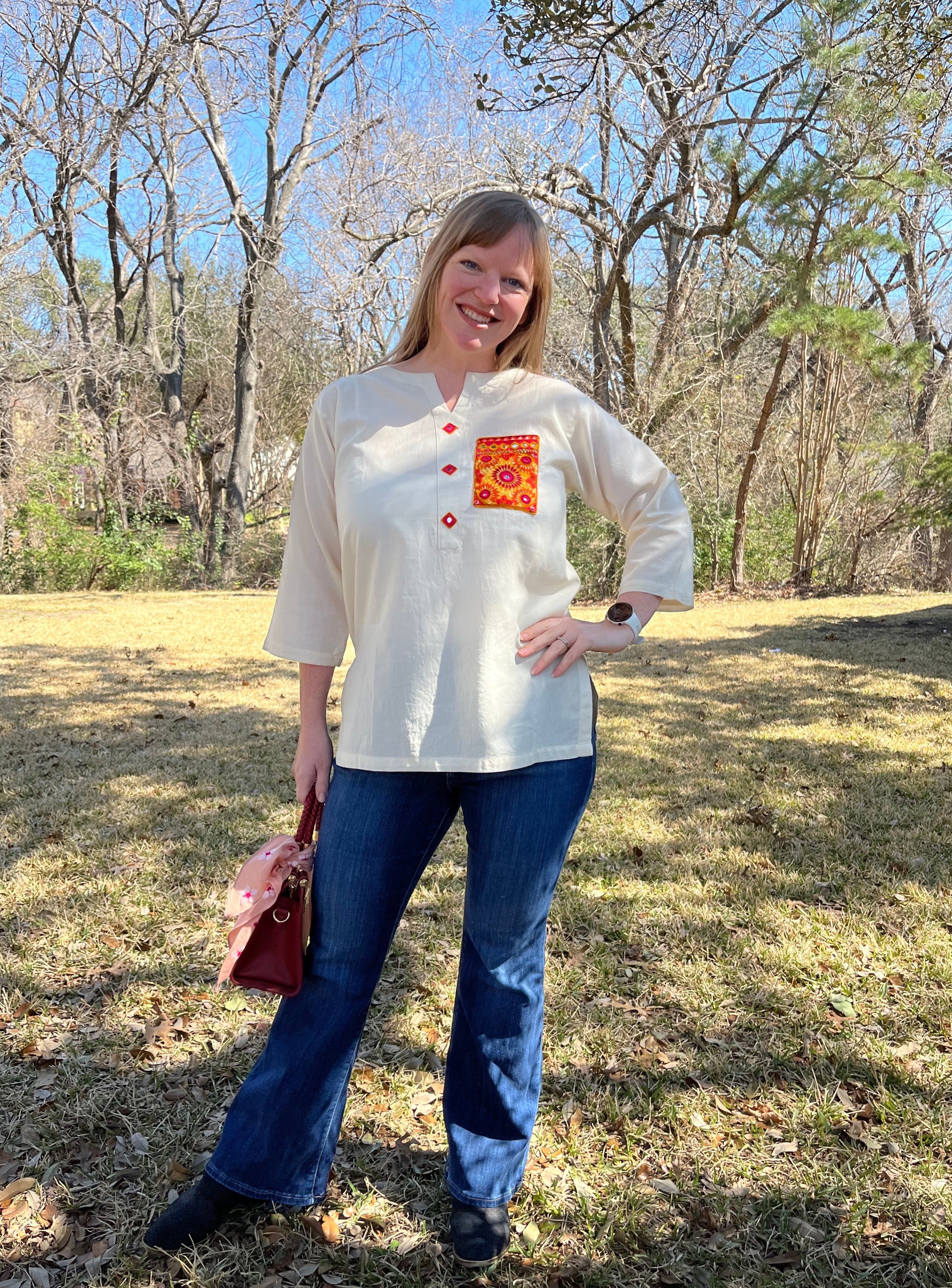 A person standing outdoors wearing a beige long-sleeved shirt with red hand-embroidered details on the pocket and sleeves, paired with blue jeans and black shoes, holding a red purse.