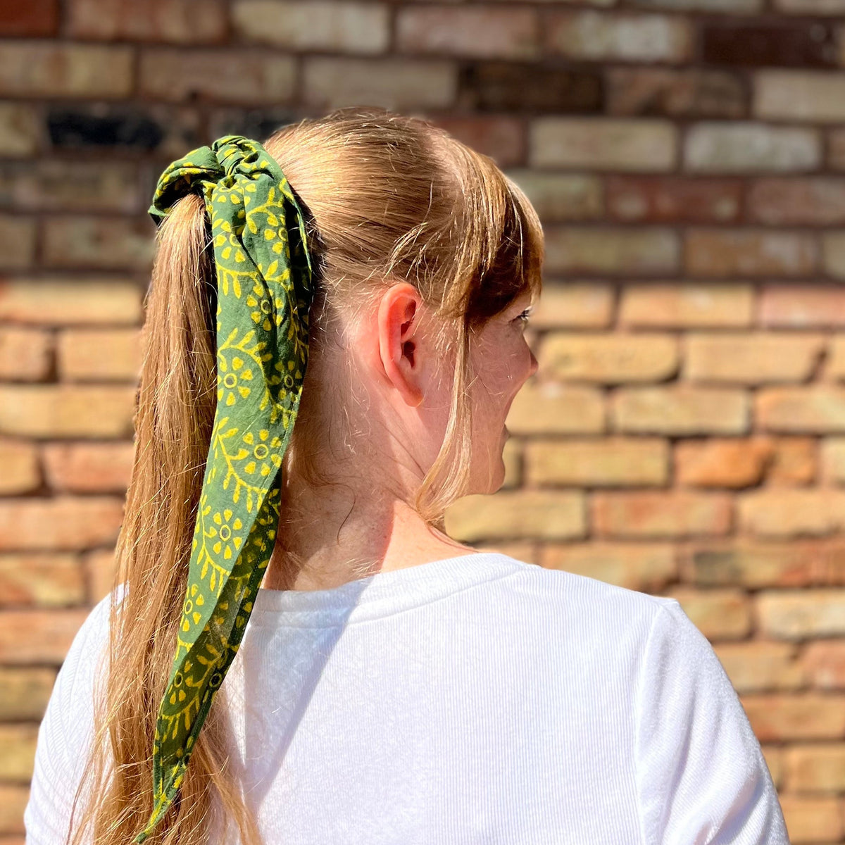 Woman with a green patterned headscarf tied in a ponytail against a brick wall.