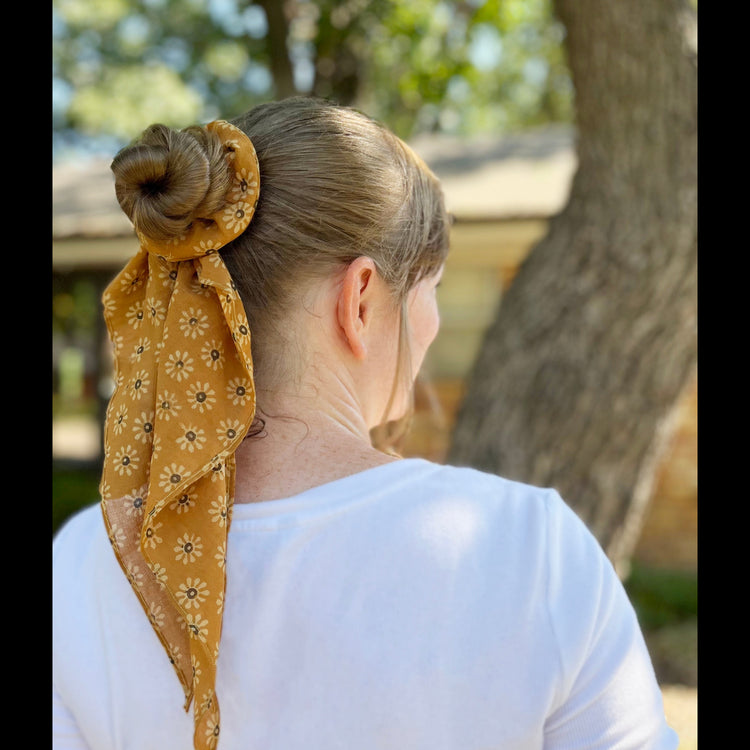 Person with a yellow blockprinted scarf tied around their hair outdoors.