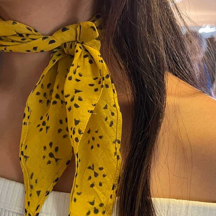 A close-up of a woman wearing a bright yellow scarf tied around her neck, featuring a black tree spot pattern.