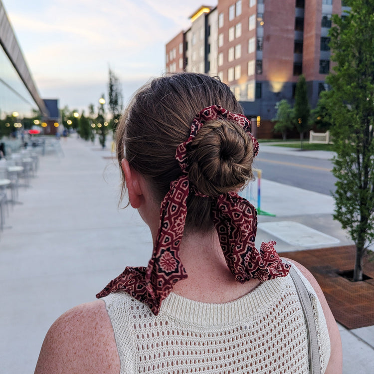 A woman viewed from behind wearing a red and black patterned scarf tied around her hair in a bun, set against an outdoor urban background with a brick building and trees.